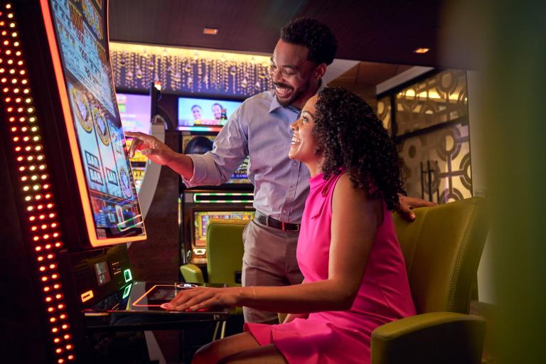 A man stands next to a seated woman at a slot machine, both are smiling