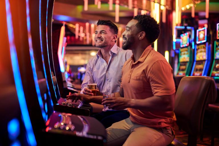 Two guests smiling and enjoying beers while playing in a vibrant, colorful casino setting.