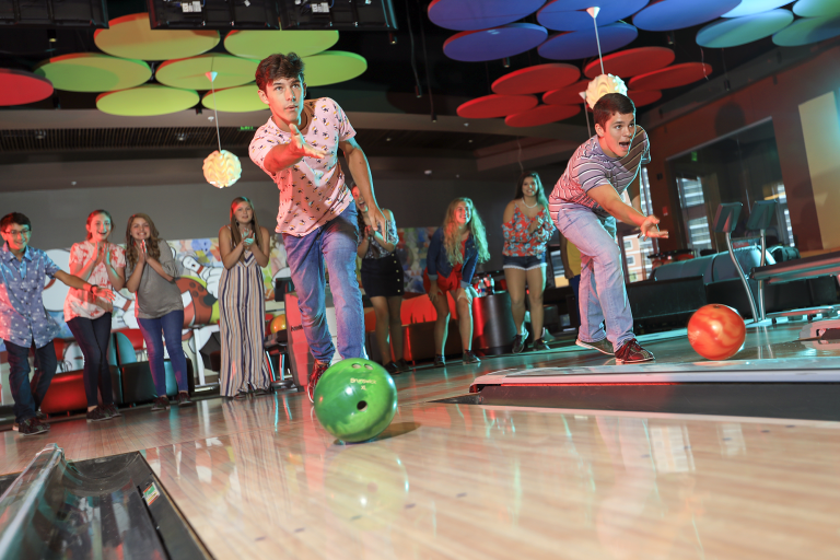 Two teen bows bowling while a group of teens watch and cheer