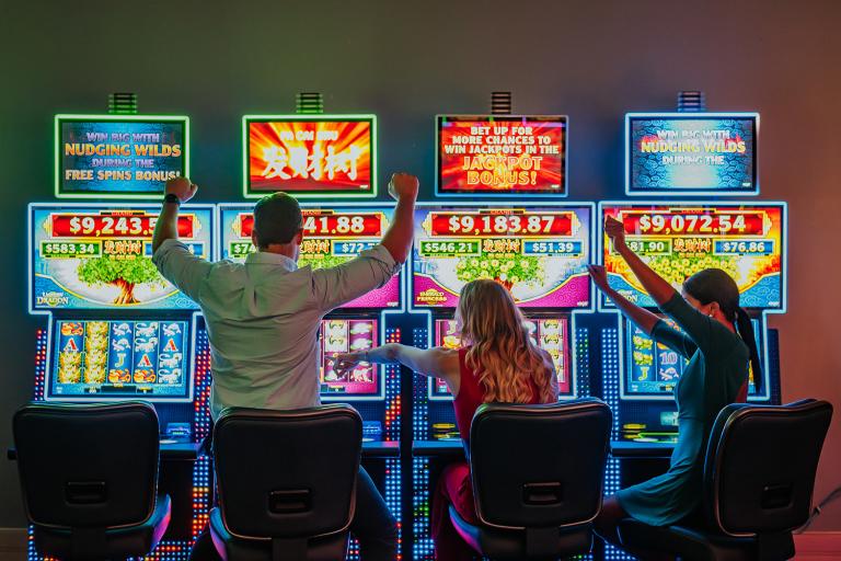 Three people sitting at bright slot machines in a casino, excitedly interacting with the games which display colorful lights and jackpot amounts.
