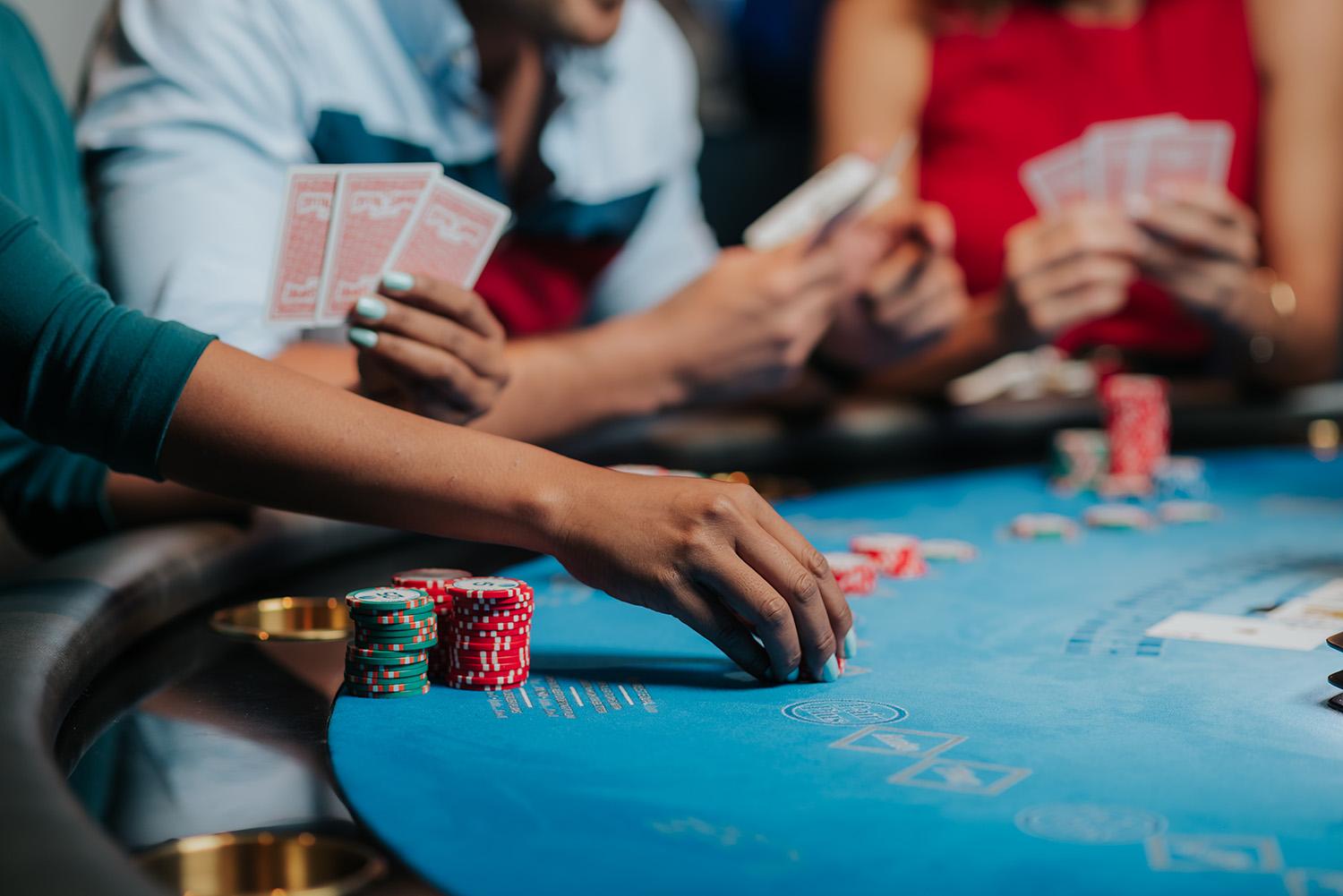 Close-up of a hand placing betting chips on a casino table with other players holding playing cards in the background, implying a tense poker game.