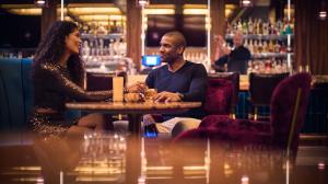 A couple has drinks at a cocktail table while a bartender shakes up another round in the background