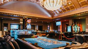 Elegant casino interior featuring a craps table surrounded by empty chairs, large chandeliers overhead, and multiple screens displaying game information.